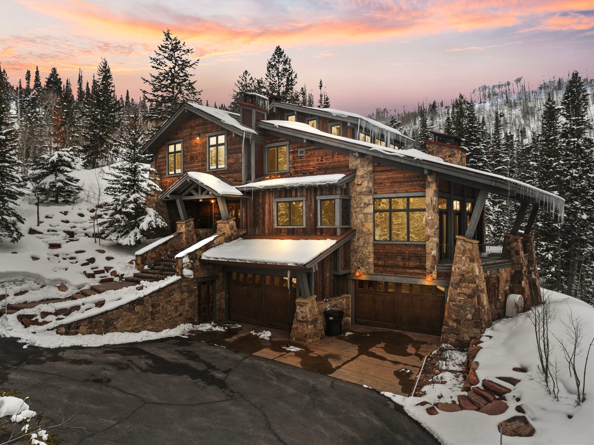 A view of Silver Horse at The Colony in Park City. The home is sitting next to a ski slope, and the windows are glowing with light. Snow covered trees surround it and there is a view of the mountain and sunset in in the background
